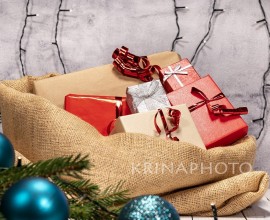Jute sack full of Christmas gifts on a white wooden table. Gifts with red, brown and silver paper with red ribbon, Christmas decorations around and twinkling lights in the background.