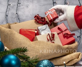 Santa Claus prepares the jute sack with Christmas gifts. Hand with white glove holding a gift box with red paper and white ribbon.