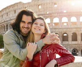Young couple traveling to Rome. The couple smiles and hugs for a selfie in front of the Colosseum.