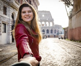 Beautiful blonde girl holds her boyfriend by the hand as he walks through the streets of the historic center of Rome. The smiling woman has long hair and is wearing a red leather jacket. In the background the Colosseum. Travel concept to Italy.
