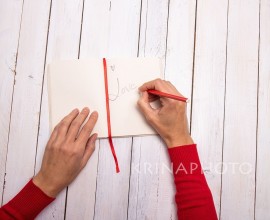 Female hands writing a lovely message on her notebook. The touch of red from the sweater and the pencil recall the Valentine color. White wooden background with empty space for text.