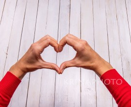 Female hands making a heart shape on a white wooden background. A touch of red from the sweater draws attention. Empty space for Valentine text.