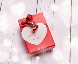 Red box with a big heart-shaped card for a special Valentine gift. White wooden background and bokeh effect all around.